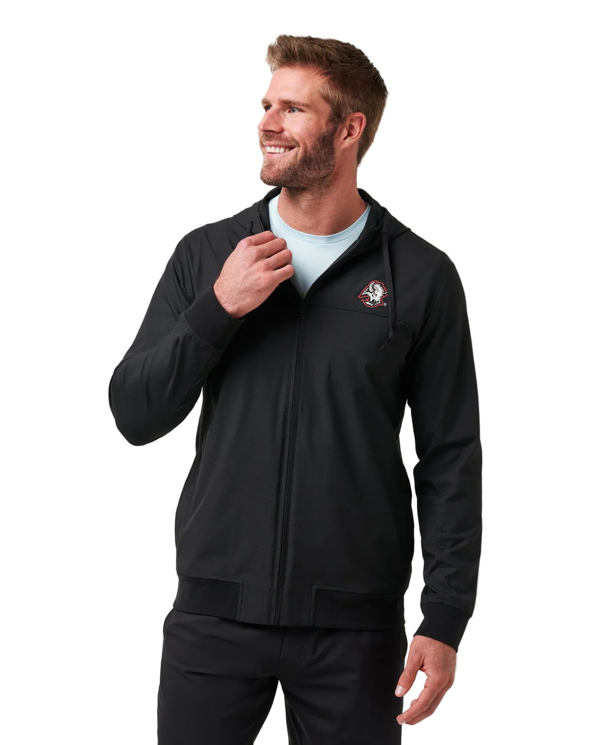 A smiling man wearing a black zip-up hoodie jacket over a light blue t-shirt and black pants, standing against a plain light gray studio background.