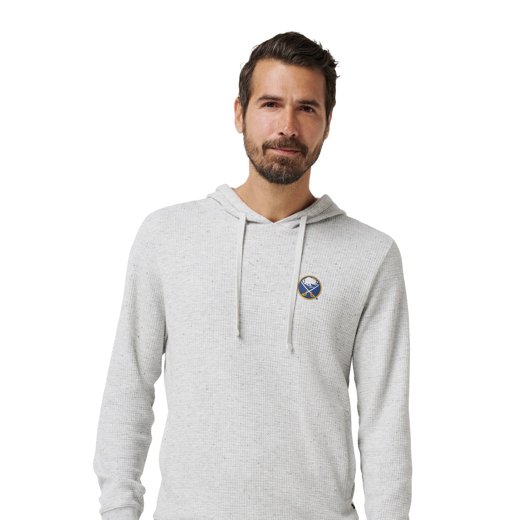A man wearing a light heather gray pullover hoodie with a small circular blue and white graphic on the left chest, paired with dark blue jeans, against a plain white background.