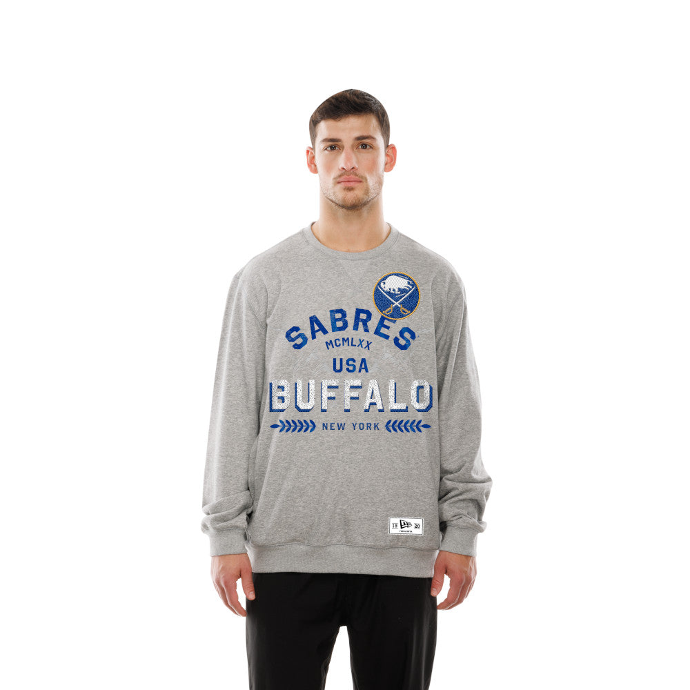 A man modeling a heather gray crewneck sweatshirt with a blue and white Buffalo Sabres graphic on the chest, paired with black pants against a solid white background.