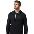 A man wearing a black pullover hoodie with cream drawstrings and a small embroidered logo on the left chest, paired with gray pants, against a plain white background.
