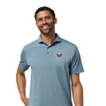 A man wearing a slate blue short-sleeve polo shirt with a small circular embroidered logo on the left chest, paired with black pants, photographed against a plain white background.