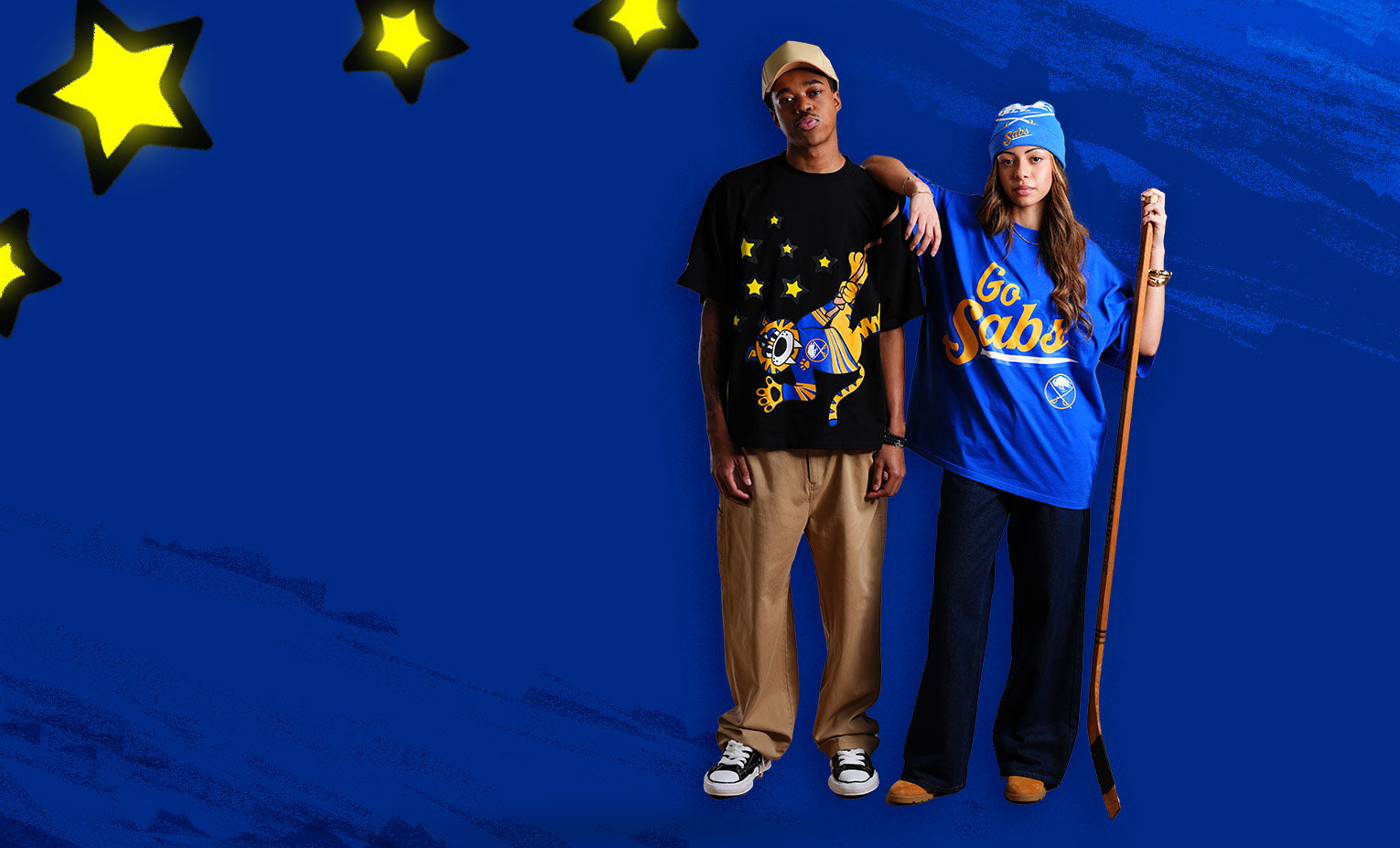 Two people standing against a blue background with yellow stars, one wearing a black t-shirt and beige pants, the other in a blue 'Go Gabi' shirt and dark pants.